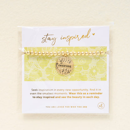 The Darling Effect’s Stay Inspired Beaded Charm Bracelet features gold beads and a round “INSPIRE” charm, displayed on a card reading “stay inspired” with a light floral background—an uplifting, meaningful gift.