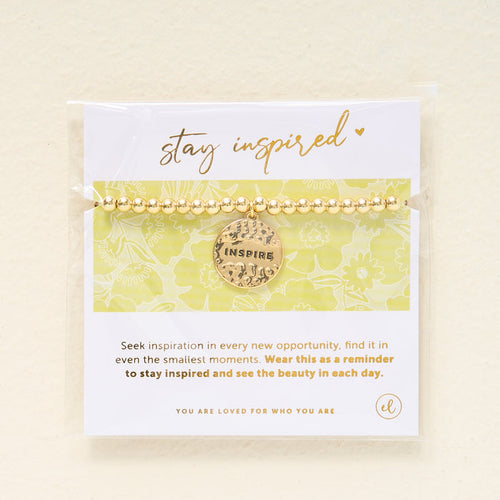 The Darling Effect’s Stay Inspired Beaded Charm Bracelet features gold beads and a round “INSPIRE” charm, displayed on a card reading “stay inspired” with a light floral background—an uplifting, meaningful gift.