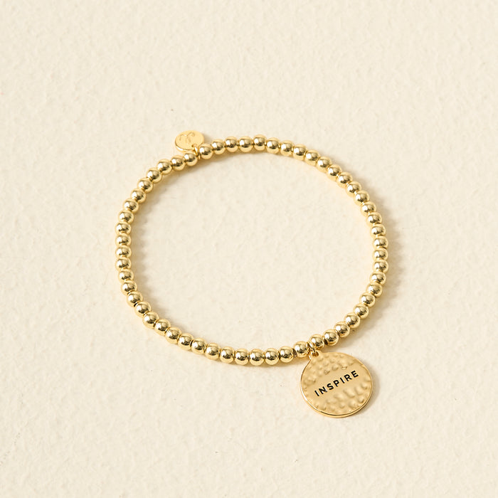 The Darling Effect’s Stay Inspired Beaded Charm Bracelet features gold beads and a round hammered “INSPIRE” charm, displayed on a light beige textured surface—a thoughtful gift for someone special.