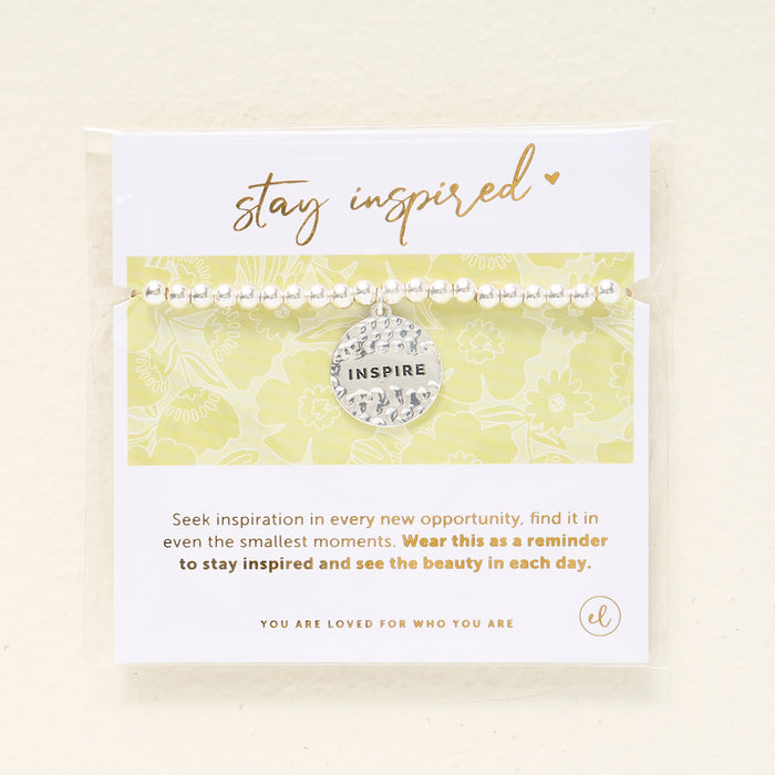 The Darling Effect's Stay Inspired Beaded Charm Bracelet features a hammered INSPIRE charm and comes on a card reading "stay inspired"—a meaningful gift to remind you to seek inspiration and appreciate the beauty each day brings.