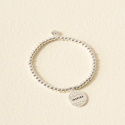 The Darling Effect's Stay Inspired Beaded Charm Bracelet features silver beads and a round charm engraved with "INSPIRE," making it a thoughtful gift. Shown on a cream-colored textured background.