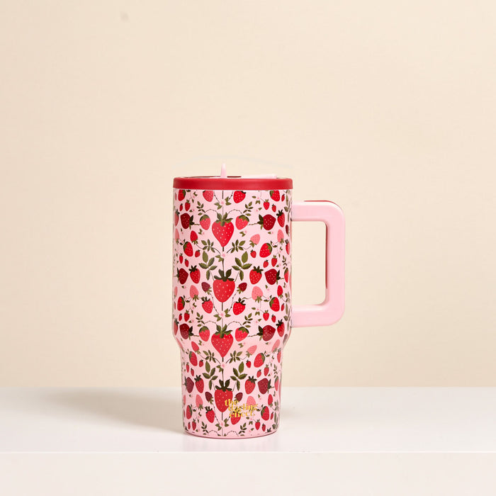 The Darling Effect's Strawberry Fields 20 oz Lifestyle Flip Straw Tumbler—a pink insulated stainless steel tumbler with a handle, decorated with strawberries and small flowers—sits on a white surface against a beige background.