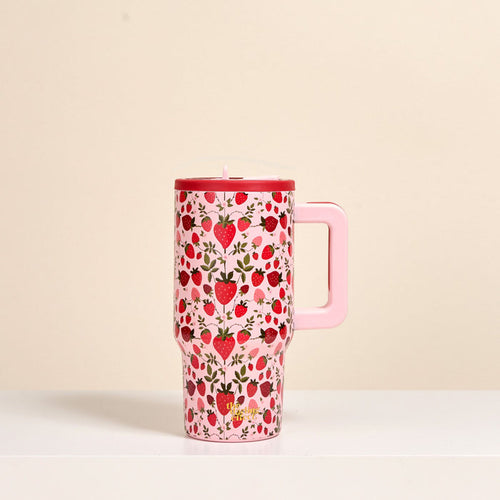 The Darling Effect's Strawberry Fields 20 oz Lifestyle Flip Straw Tumbler—a pink insulated stainless steel tumbler with a handle, decorated with strawberries and small flowers—sits on a white surface against a beige background.