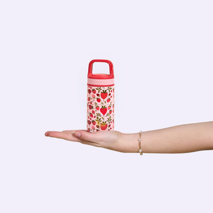 A hand with a bracelet holds The Darling Effect’s Strawberry Fields 6 oz Lil' Bitty Bottle—a petite pink water bottle adorned with red strawberries and green leaves—perfect for hydration on the go against a light background.
