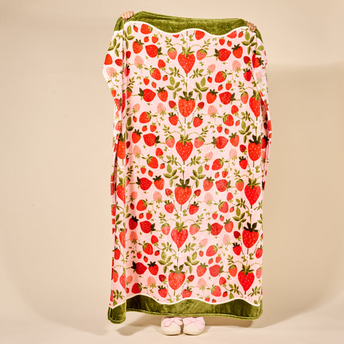 A person holds up The Darling Effect's Strawberry Fields Snuggle Up Plush Blanket, featuring a vibrant strawberry pattern. Only their hands and pink-slippered feet peek out from behind the soft blanket, set against a plain beige background.