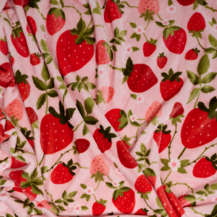 The Darling Effect’s Strawberry Fields Snuggle Up Plush Blanket features a pink background with red and pink strawberries and green leaves, offering a soft, beautifully draped, slightly wrinkled plush fabric.