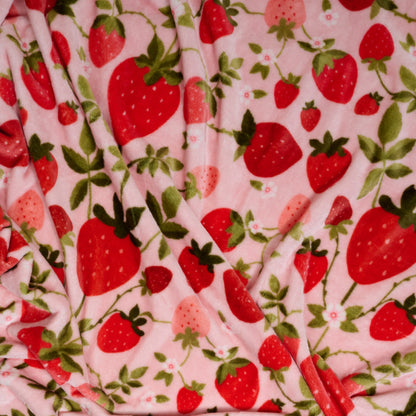 The Darling Effect’s Strawberry Fields Snuggle Up Plush Blanket features a pink background with red and pink strawberries and green leaves, offering a soft, beautifully draped, slightly wrinkled plush fabric.