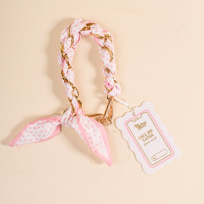 The Darling Effect Sugar Dot Call Me Later Phone Scarf: a chic pink and white polka dot braided wrist strap with gold-tone chain, attached to a pink-edged tag, displayed on a beige surface.