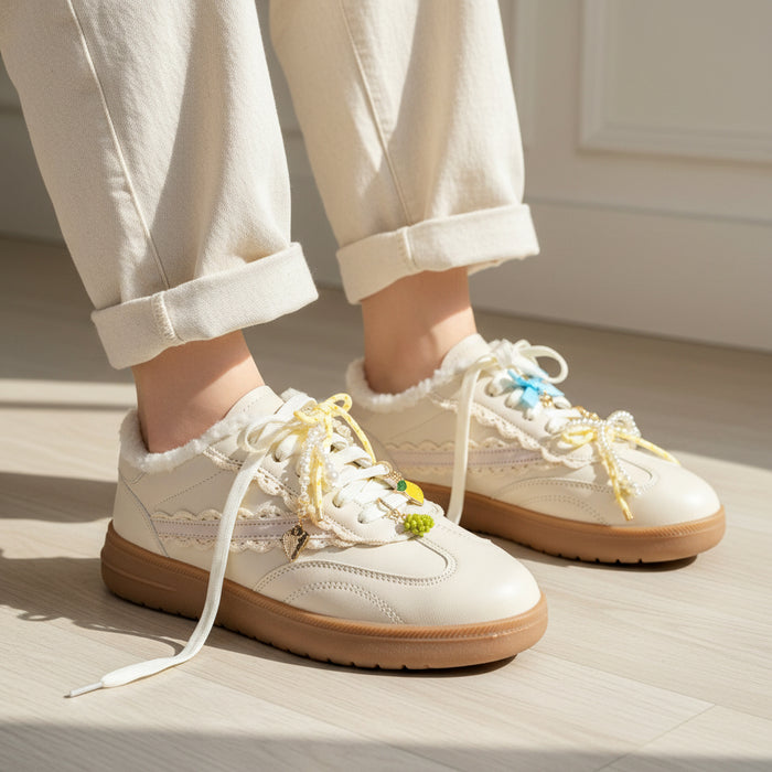 A person wears cream lace-up sneakers with tan soles and The Darling Effect’s Summer Soirée Dazzle Shoe Charm Pack, paired with light cuffed pants, standing on a light wood floor in bright natural light.