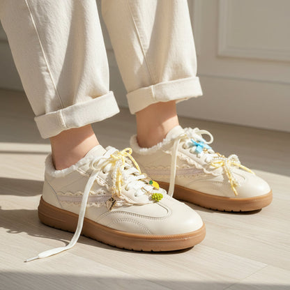 A person wears cream lace-up sneakers with tan soles and The Darling Effect’s Summer Soirée Dazzle Shoe Charm Pack, paired with light cuffed pants, standing on a light wood floor in bright natural light.