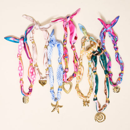 The Darling Effect's Sunlit Bloom Make a Statement Scarf Necklace features colorful silk with gold bead accents and a unique gold charm pendant, offering artful patterns on a cream background for standout style.