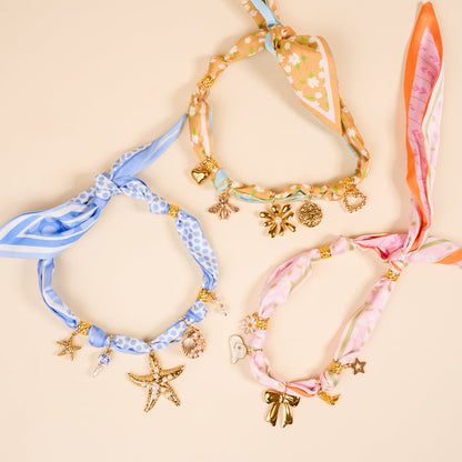 Three Sunlit Bloom Make a Statement Scarf Necklaces by The Darling Effect—blue, peach, and pink—form a circle. Each displays unique patterns and gold star, seashell, bow, and heart charms on a pale beige background.