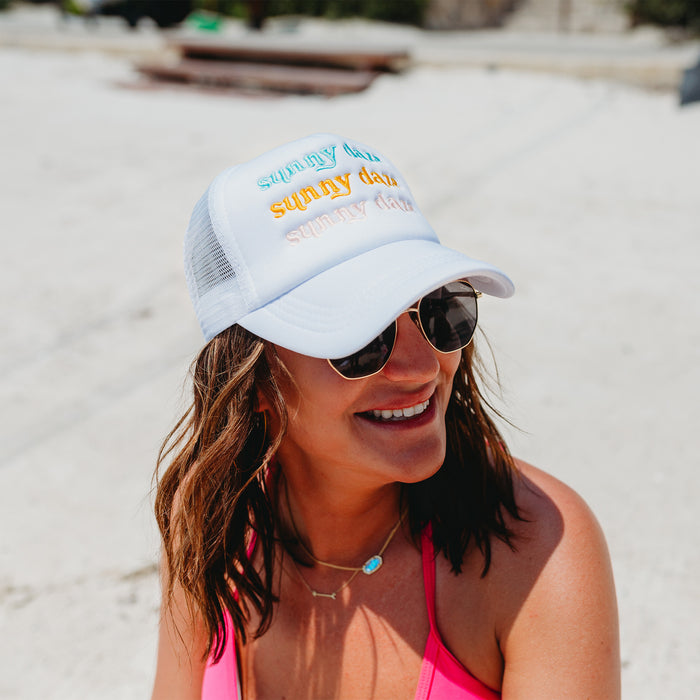 A woman in sunglasses and a pink swimsuit top smiles on a sunny beach while wearing the-darling-effect's Sunny Daze Trucker Hat, featuring colorful "sunny days" embroidery.