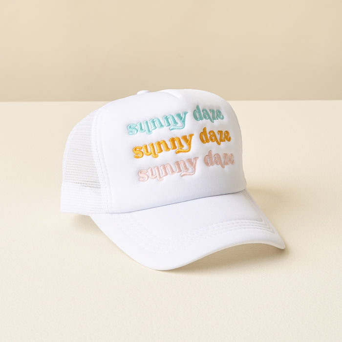 The-darling-effect’s Sunny Daze Trucker Hat features “sunny daze” embroidered three times in pastel blue, yellow, and pink on white mesh, shown atop a cream surface with a beige background.