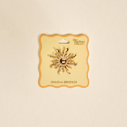 The Sunshine Timeless Brooch by The Darling Effect, a vintage-inspired gold sun-shaped piece with wavy rays, is displayed on a cream card labeled "the darling effect" and "timeless BROOCH" against a neutral background.