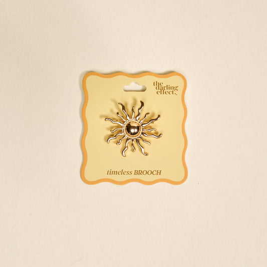 The Sunshine Timeless Brooch by The Darling Effect, a vintage-inspired gold sun-shaped piece with wavy rays, is displayed on a cream card labeled "the darling effect" and "timeless BROOCH" against a neutral background.