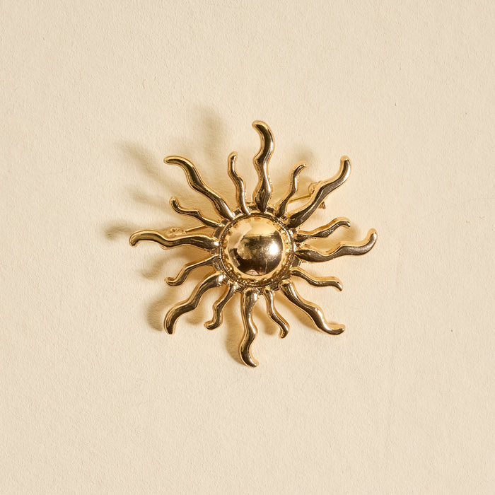 The Darling Effect's Sunshine Timeless Brooch is a gold, sun-shaped vintage-inspired accessory with wavy rays and a round center, beautifully showcased against a light beige background.