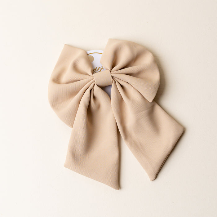 A large Tan Hair Bow by The Darling Effect sits on a cream background, its label partially visible. This timeless accessory brings a touch of elegance to any outfit.