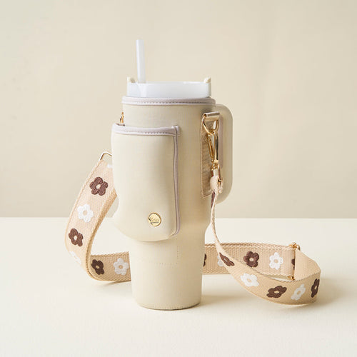 The Darling Effect's Tan Tumbler Sling features a beige insulated carrier with an adjustable strap, handle, detachable pouch, and brown and white floral accents. It holds a white cup with lid and straw, shown against a light background.