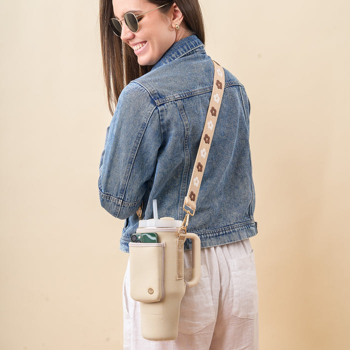 A woman in sunglasses and a denim jacket smiles, carrying The Darling Effect’s Tan Tumbler Sling—a large beige neoprene carrier with an adjustable strap, phone pocket, and floral pattern that completes her stylish look.