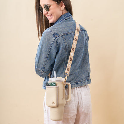 A woman in sunglasses and a denim jacket smiles, carrying The Darling Effect’s Tan Tumbler Sling—a large beige neoprene carrier with an adjustable strap, phone pocket, and floral pattern that completes her stylish look.