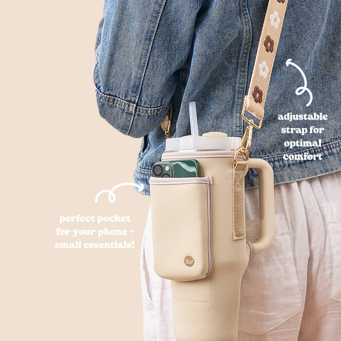 A person in a denim jacket and light pants carries The Darling Effect’s Tan Tumbler Sling, a neoprene tumbler carrier with an adjustable strap, beige color, phone side pocket, and text emphasizing comfort and utility.