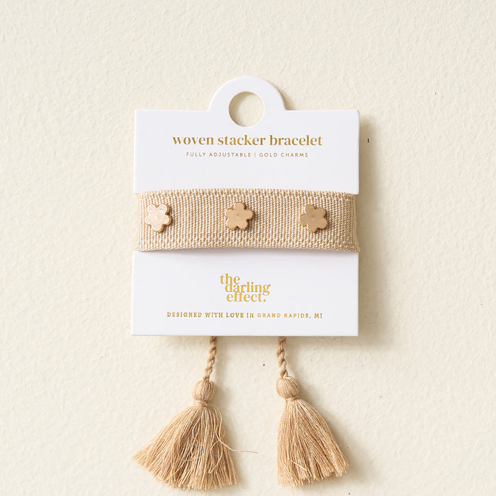 A Tan Woven Bracelet by The Darling Effect, featuring gold flower charms and adjustable tassel ends, is presented on a white card labeled "woven stacker bracelet.