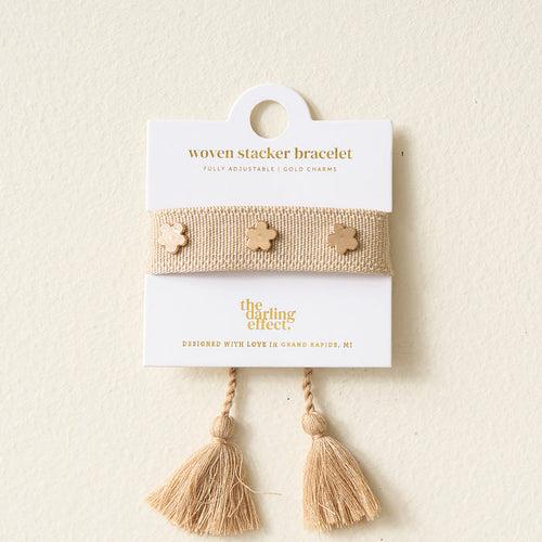 A Tan Woven Bracelet by The Darling Effect, featuring gold flower charms and adjustable tassel ends, is presented on a white card labeled "woven stacker bracelet.