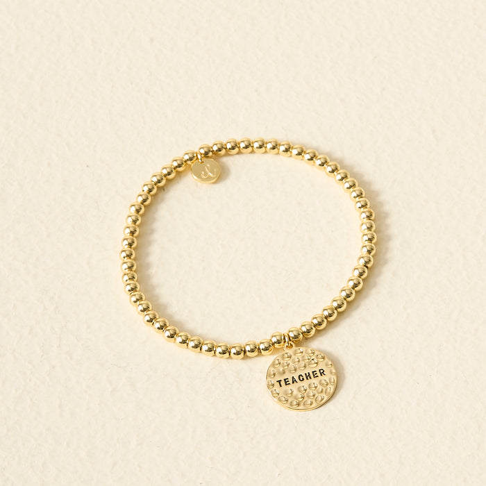 The Darling Effect's Teacher Beaded Charm Bracelet features gold beads and a round sentiment charm engraved with "TEACHER," shown on a light cream surface—the perfect thoughtful gift for educators.