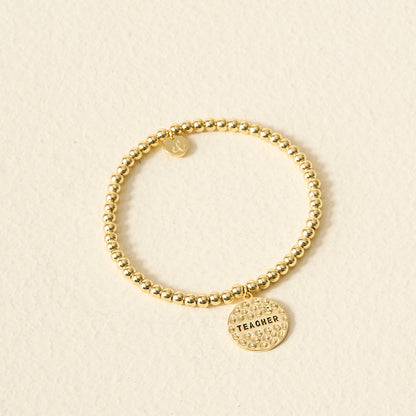 The Darling Effect's Teacher Beaded Charm Bracelet features gold beads and a round sentiment charm engraved with "TEACHER," shown on a light cream surface—the perfect thoughtful gift for educators.