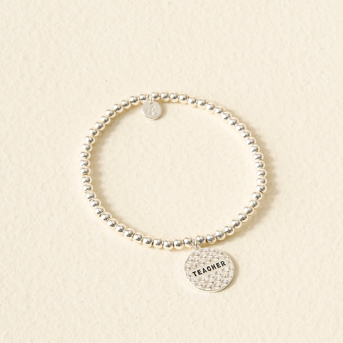 The Darling Effect’s Teacher Beaded Charm Bracelet features a round, textured "TEACHER" charm—an ideal, thoughtful gift and perfect Sentiment Charm Bracelet for educators, displayed on a light beige background.