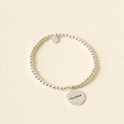 The Darling Effect’s Teacher Beaded Charm Bracelet features a round, textured "TEACHER" charm—an ideal, thoughtful gift and perfect Sentiment Charm Bracelet for educators, displayed on a light beige background.