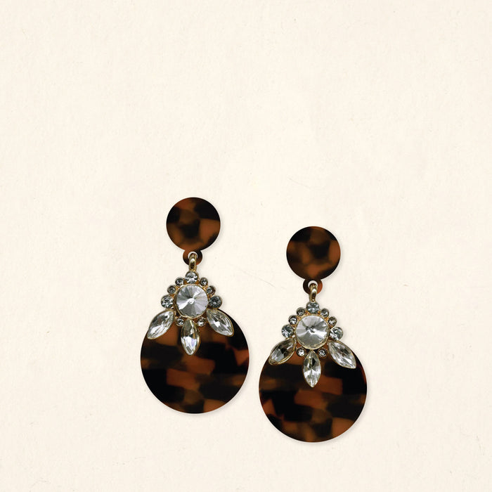 The Darling Effect’s Totally Tortoise Show Stopper Earrings feature dark tortoiseshell round disks with clear rhinestones and marquise gems in a floral pattern on a light beige background.