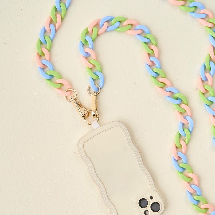 The Darling Effect Tri Lively Hold the Phone Crossbody Chain in pastel pink, green, and blue is paired with a beige wavy-edged phone case, creating a chic accessory set shown on a light cream surface.