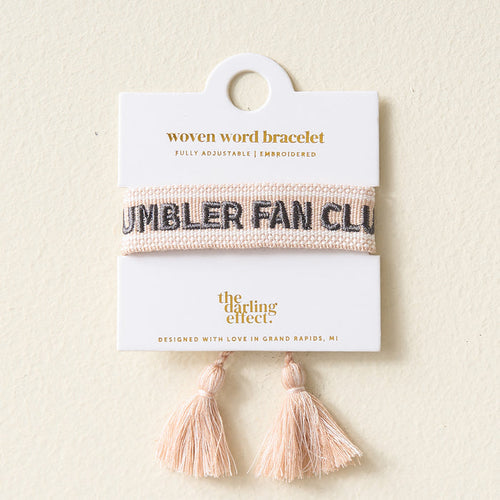 The Darling Effect’s Tumbler Fan Club Woven Bracelet is a trendy, adjustable piece featuring “TUMBLER FAN CLUB” in black, two pink tassels, and comes displayed on a branded card.
