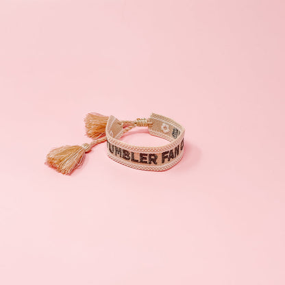 The Darling Effect's Tumbler Fan Club Woven Bracelet features trendy embroidery, playful tassels, and an adjustable fit—perfect for adding style to any outfit. Shown against a pink background.