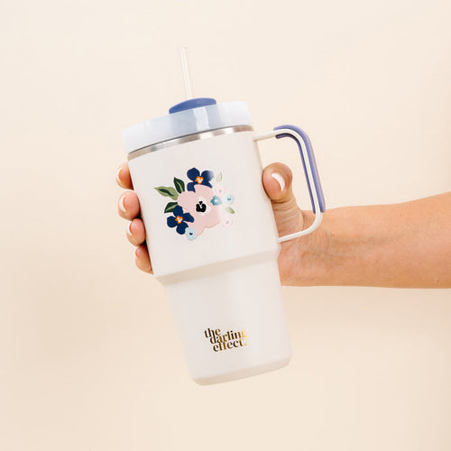 A hand holds The Darling Effect uds & Blooms Cream 20oz Mini On-the-Go Tumbler with a blue handle, floral and cloud design, blue lid, reusable straw, and “the darling effect” in gold near the bottom.