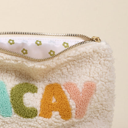 The Darling Effect’s Vacay Teddy Pouch is a fuzzy cream sherpa pouch with a gold zipper, “CAY” embroidered in green, yellow, and pink, and a white lining printed with tiny green flowers—a cute addition to your teddy pouches set.