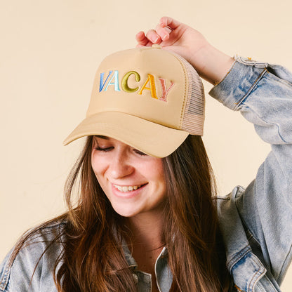 A young woman in a denim jacket sports the-darling-effect's Vacay Trucker Hat, featuring "VACAY" in colorful embroidery—one of this season's trendiest picks.