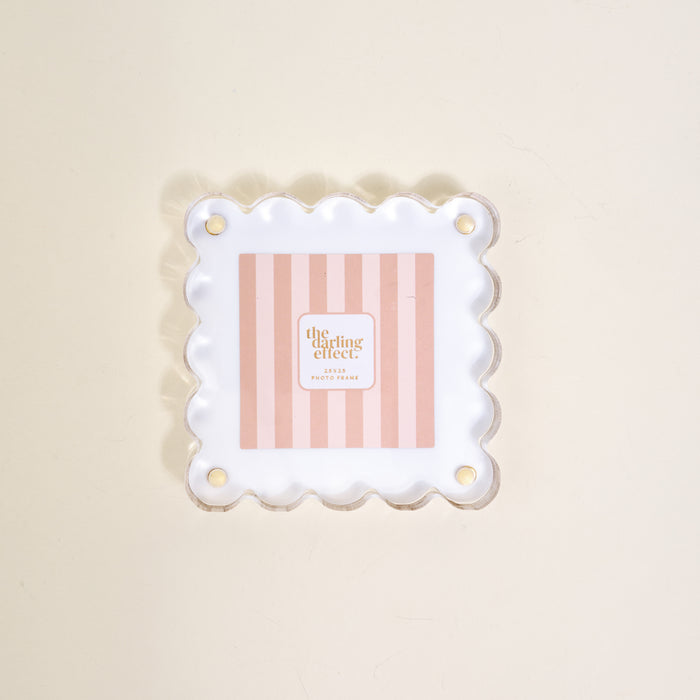 The Darling Effect's "White Little Moments Acrylic Picture Frame" showcases a square acrylic frame with scalloped edges and gold corner holes, displayed on a light beige surface.