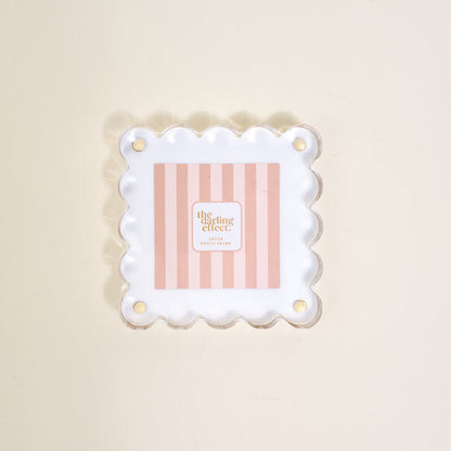 The Darling Effect's "White Little Moments Acrylic Picture Frame" showcases a square acrylic frame with scalloped edges and gold corner holes, displayed on a light beige surface.
