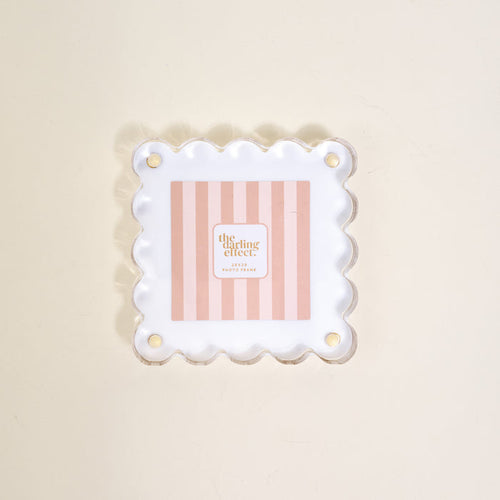 The Darling Effect's "White Little Moments Acrylic Picture Frame" showcases a square acrylic frame with scalloped edges and gold corner holes, displayed on a light beige surface.
