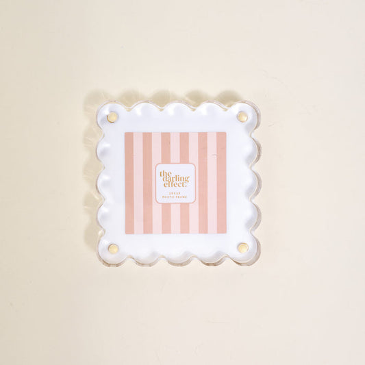 The Darling Effect's "White Little Moments Acrylic Picture Frame" showcases a square acrylic frame with scalloped edges and gold corner holes, displayed on a light beige surface.