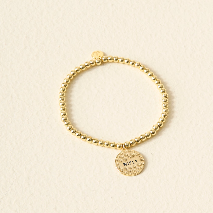 The Darling Effect’s Wifey Beaded Charm Bracelet features gold beads and a round, textured charm engraved with "WIFEY." Displayed on a light beige background, it’s a beautiful and meaningful gift.