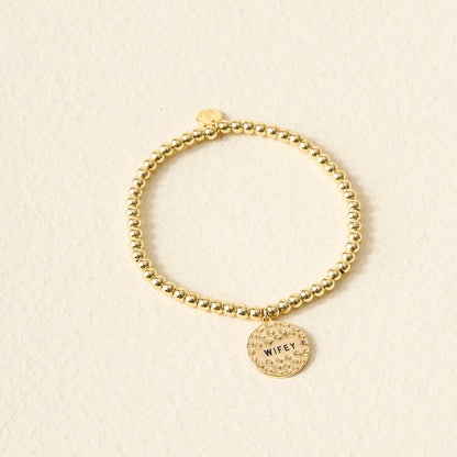 The Darling Effect’s Wifey Beaded Charm Bracelet features gold beads and a round, textured charm engraved with "WIFEY." Displayed on a light beige background, it’s a beautiful and meaningful gift.