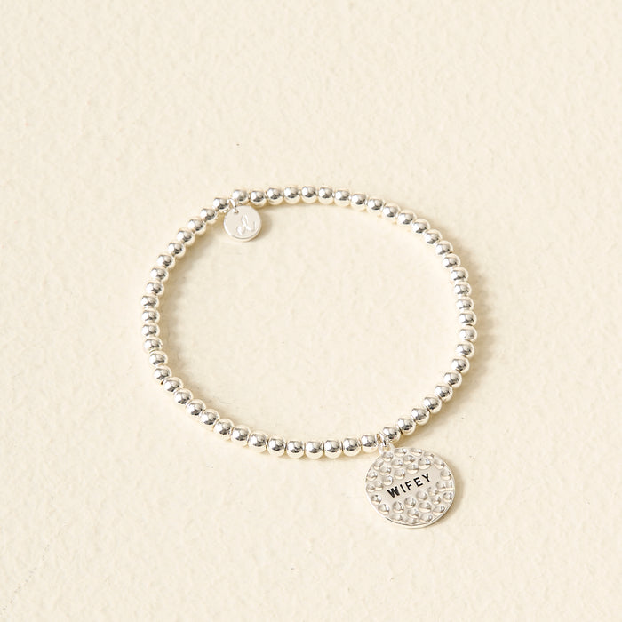 The Darling Effect’s Wifey Beaded Charm Bracelet features two round charms, including one engraved with "WIFEY," on a silver beaded band—displayed on a cream textured background, it makes a meaningful gift and stylish accessory.