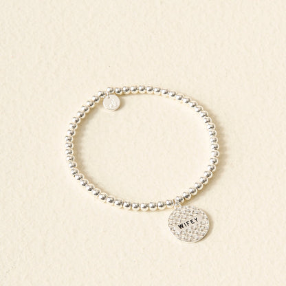 The Darling Effect’s Wifey Beaded Charm Bracelet features two round charms, including one engraved with "WIFEY," on a silver beaded band—displayed on a cream textured background, it makes a meaningful gift and stylish accessory.