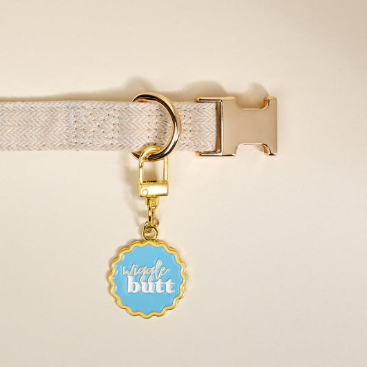 Add charm to your pet’s style with The Darling Effect’s Wiggle Butt Pawfect Collar Charm—a round, gold-edged blue tag with white and yellow "wiggle butt" text—perfect for any beige fabric dog collar with a gold buckle.