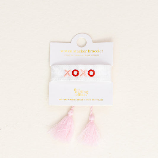 The XOXO Woven Bracelet by the-darling-effect features white woven fabric, pink and red embroidered "XOXO," two light pink tassels, and comes on a white card with gold text.