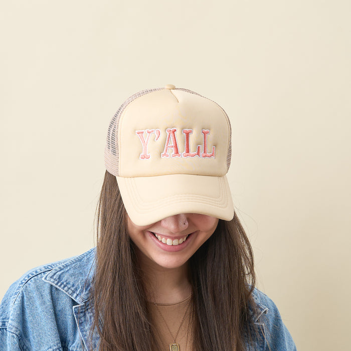 A person with long brown hair smiles and looks down, wearing the-darling-effect's Y'all Trucker Hat—beige with "YALL" in pink—and a blue denim jacket, standing against a light beige background.